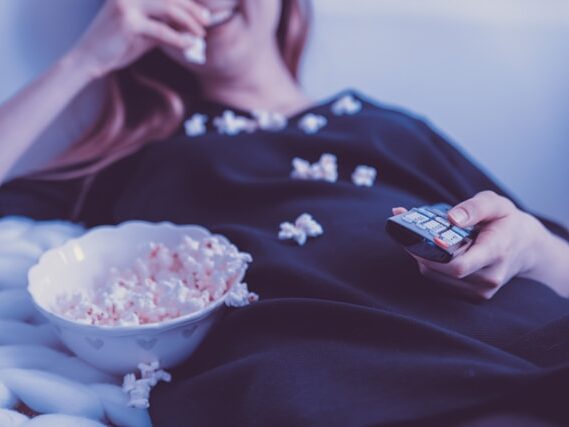 女性が寝そべりながらポップコーンを食べてリモコンを握っている画像